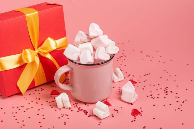 Uma caneca rosa está deitada de lado com marshmallows polvilhados em forma de coração e presente