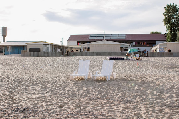 Uma cena de praia com duas cadeiras brancas na areia e um prédio ao fundo.