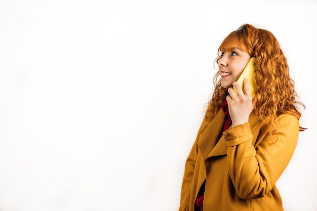 Uma cena média de uma mulher ruiva feliz falando ao telefone com um casaco amarelo em uma parede branca isolada