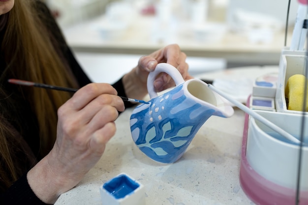 Foto uma ceramista irreconhecível pinta um leiteiro de barro com um pincel