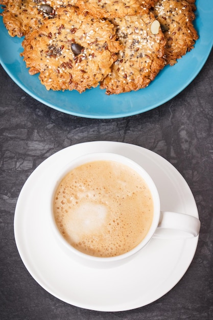 Uma chávena de café branco e biscoitos de aveia caseiros frescos cozidos com mel e diferentes sementes saudáveis em prato azul Deliciosa sobremesa crocante