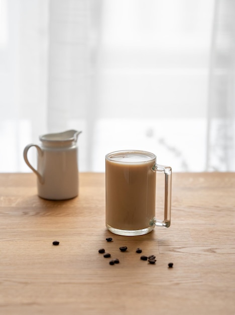 Uma chávena de café laltte com leite em uma mesa de madeira com grãos de café e jarro de leite contra o fundo de uma janela