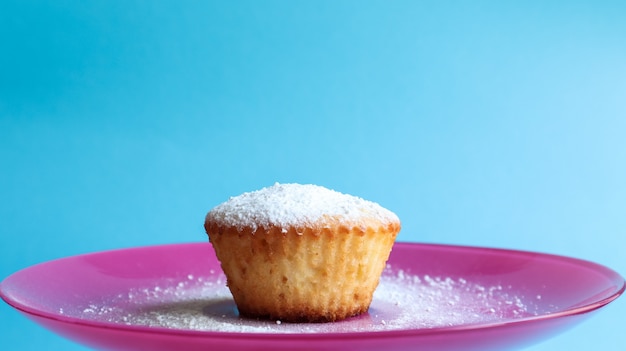 Uma coalhada polvilhada com açúcar de confeiteiro em um prato rosa, sobre um fundo azul, vista lateral.