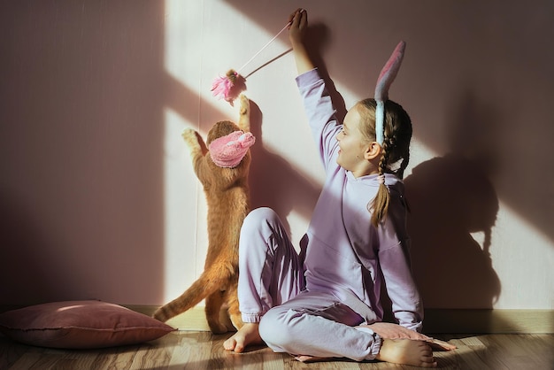 uma colegial na cabeça com orelhas de coelho brinca com um gato ruivo com um chapéu de Páscoa ao sol