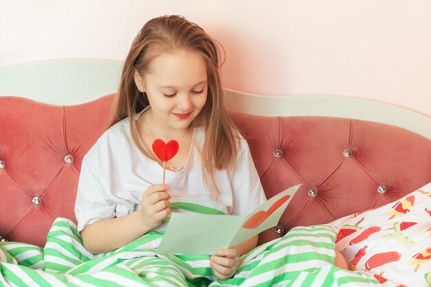 uma colegial senta-se em sua cama e lê um cartão caseiro com um coração