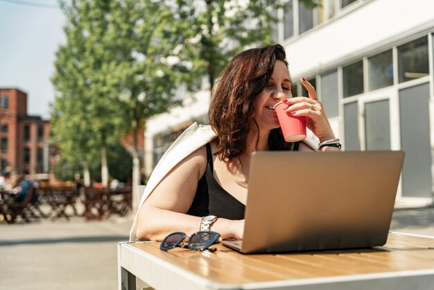 Foto uma confortável gerente feminina trabalha ao ar livre em um café usa um laptop e bebe café