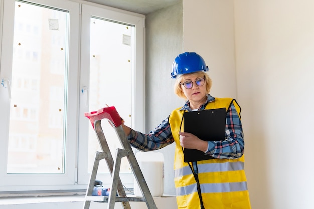 Uma Construtora com um tablet nas mãos inspeciona uma sala em um novo prédio. Mulher de meia-idade com um capacete faz reparos no apartamento.