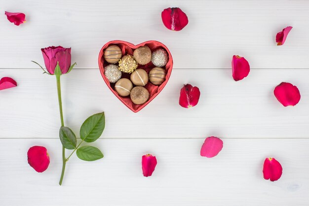 Uma costa da rosa e do chocolate do vermelho em um coração deu forma à caixa em uma tabela de madeira branca.