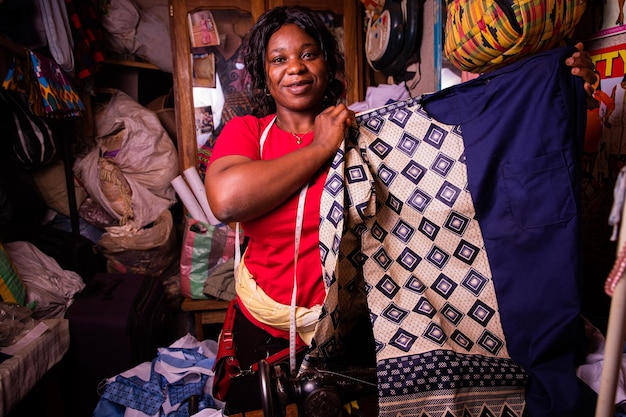 Uma costureira africana feliz mostra o vestido que acabou de costurar Terno africano típico para homens recém-feitos