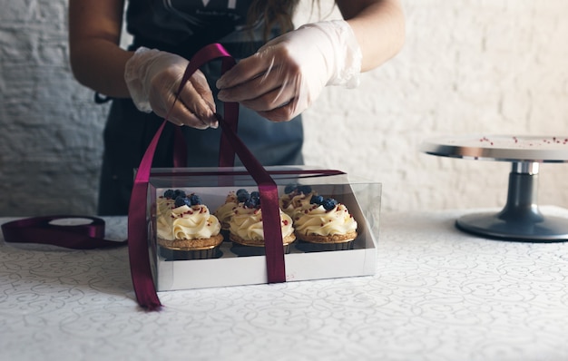 Uma cozinheira de avental cinza embala cupcakes com creme em uma caixa de presente para enviar o pedido ao cliente. assar em casa.