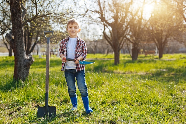 Uma criança bonita em pé com uma colher de solo no jardim de uma casa e olhando para a frente em um lindo dia ensolarado