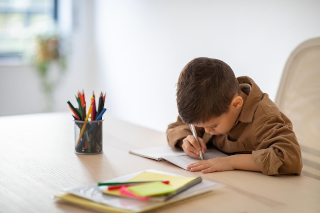 Uma criança caucasiana ocupada escreve em um caderno desenha estudos e gosta de lição de casa na mesa no interior de uma sala clara