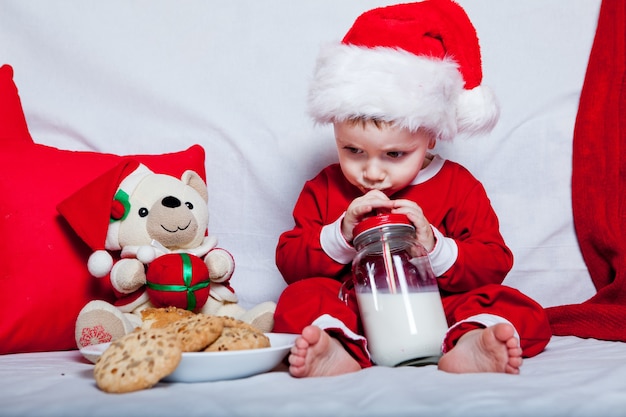 Uma criança com um chapéu de Papai Noel vermelho come biscoitos e leite. Retrato de Natal de um bebê com um chapéu de Papai Noel vermelho.