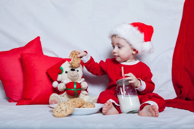 Uma criança com um chapéu de Papai Noel vermelho come biscoitos e leite. Retrato de Natal de um bebê com um chapéu de Papai Noel vermelho.