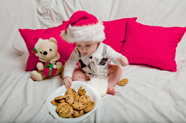 Uma criança com um chapéu de papai noel vermelho come biscoitos e leite. retrato de natal de um bebê com um chapéu de papai noel vermelho.