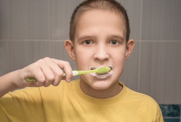 Uma criança com uma camiseta amarela escova os dentes pela manhã com uma escova de dentes. No banheiro.