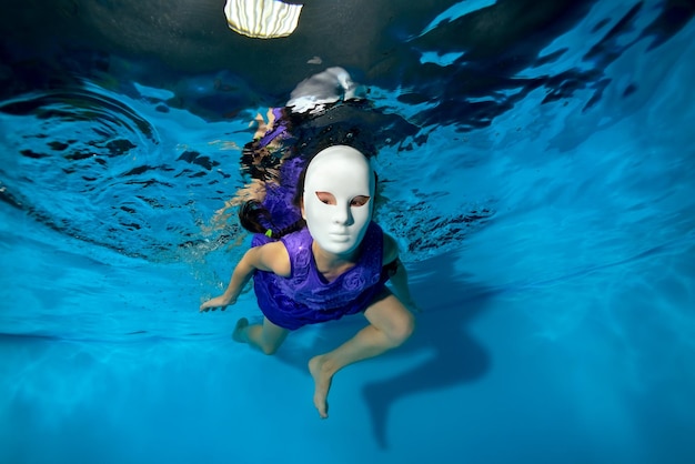 Uma criança com uma máscara branca de máscaras nada debaixo d'água na piscina e olha para a câmera