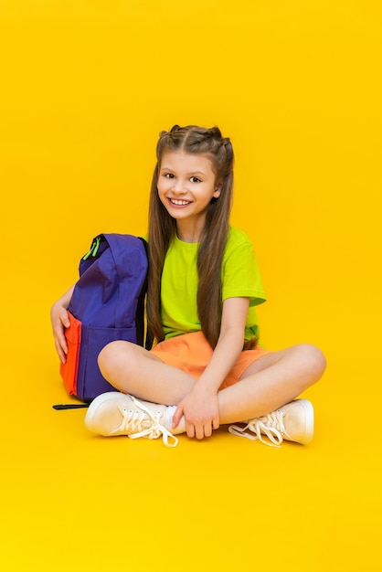 Uma criança com uma mochila escolar Uma garotinha de short está sentada e se preparando para aulas extras de verão Educação infantil Fundo amarelo isolado