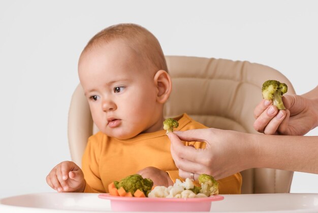 Uma criança de 11 a 15 meses se recusa a comer comida vegetariana. a mão da mãe estende brócolis cozido para a criança, a criança vira a cabeça para longe da comida