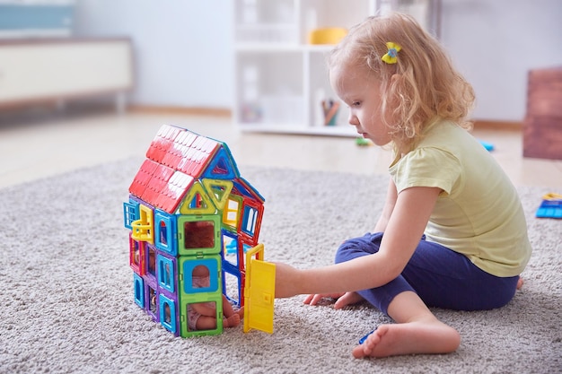 Uma criança de cabelos cacheados coleciona uma casa feita de peças de design coloridas no tapete Copiar espaço