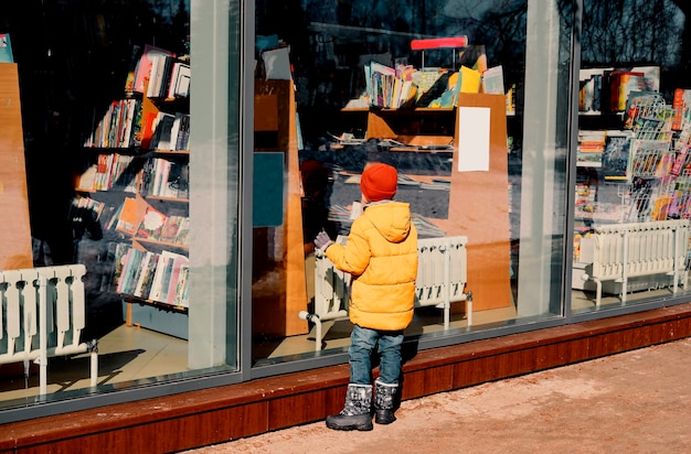Uma criança de paletó amarelo examina atentamente a janela de um quiosque de rua.