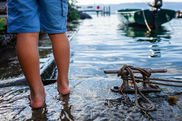 Foto uma criança de pés descalços na beira de um lago com água clara e as pedras ancorado barco e uma doca de metal