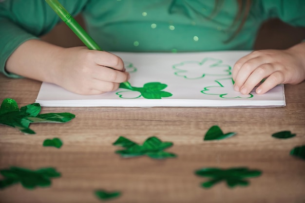 Uma criança em casa à mesa desenha e corta trevos verdes para o Dia de São Patrício