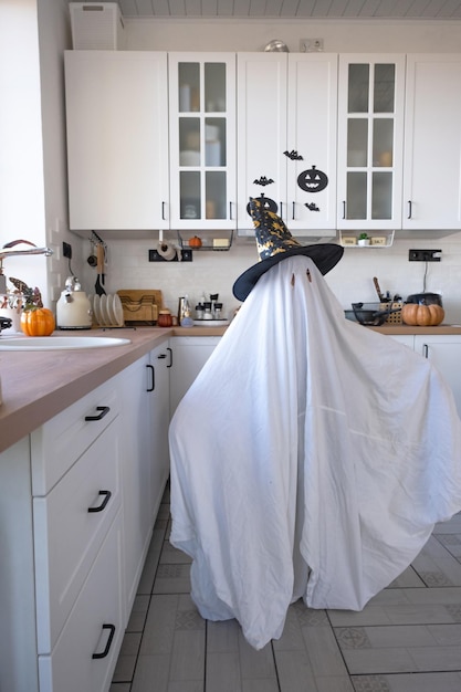 Uma criança em lençóis com recorte para olhos como uma fantasia de fantasma na cozinha decorada para o feriado de Halloween Um fantasma engraçado e engraçado Festa de Halloween