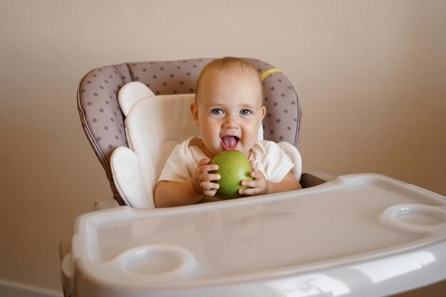 uma criança em uma cadeira infantil em uma roupa branca brinca com uma maçã verde Comida