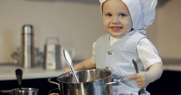Uma criança engraçada com o garfo e a colher na cozinha Retrato de um menino sujo com um chapéu culinário