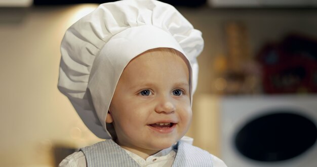 Uma criança engraçada com o garfo e a colher na cozinha Retrato de um menino sujo com um chapéu culinário