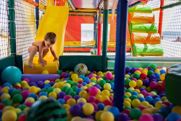 Uma criança engraçada e despreocupada brinca em uma pequena sala de jogos e rola de um pequeno escorregador brilhante em uma piscina com pequenas bolas coloridas