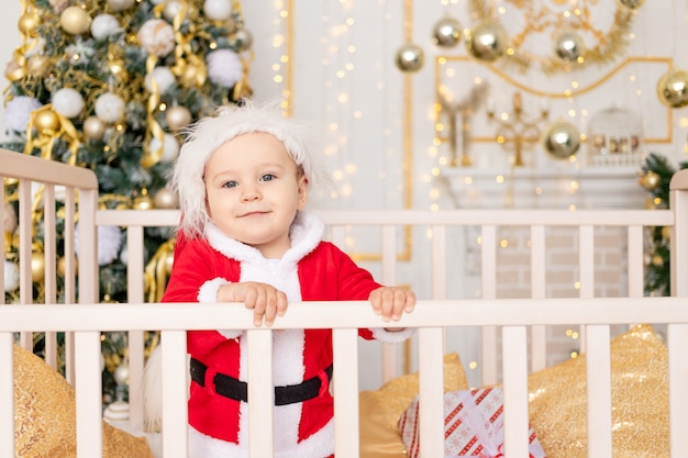 Uma criança fantasiada de Papai Noel está em um berço em casa perto de uma árvore de Natal