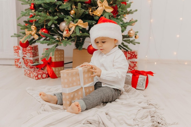 Uma criança feliz com um chapéu de Papai Noel se senta no chão perto da árvore de Natal e tem uma caixa com um presente nas mãos.