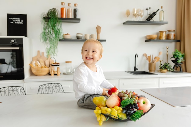 Uma criança feliz sentada na cozinha sorrindo ao lado de frutas e vegetais comem reduzidos