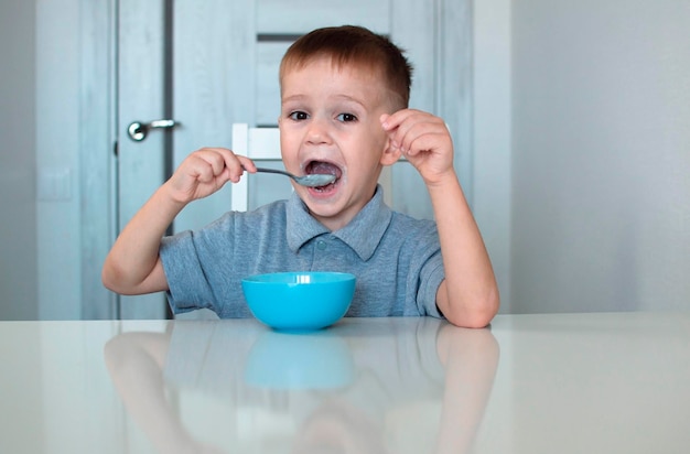 Uma criança feliz toma um café da manhã saudável e delicioso. Retrato de uma criança tomando café da manhã. Um bebê fofo e feliz está sentado na cozinha e aprendendo com uma colher. Foco seletivo.