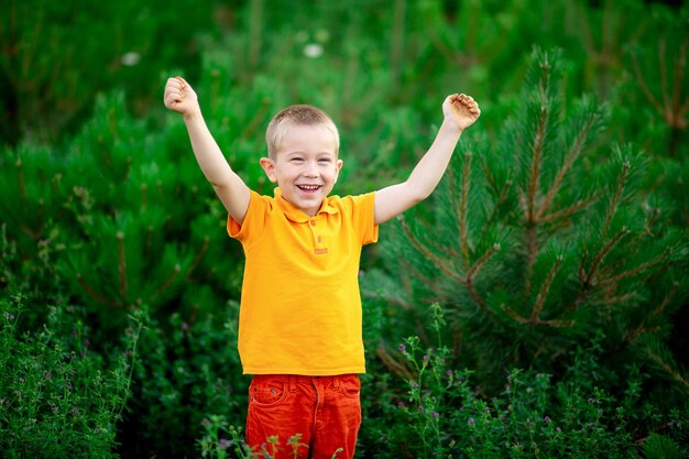 Uma criança feliz, um menino com roupas laranja fica parado na grama verde no verão e levanta as mãos