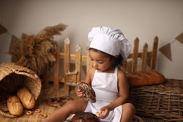 Uma criança fofa de pele escura com cabelo encaracolado e fantasiada de chef comendo um pãozinho e um pãozinho