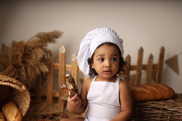 Uma criança fofa de pele escura com cabelo encaracolado e fantasiada de chef comendo um pãozinho e um pãozinho