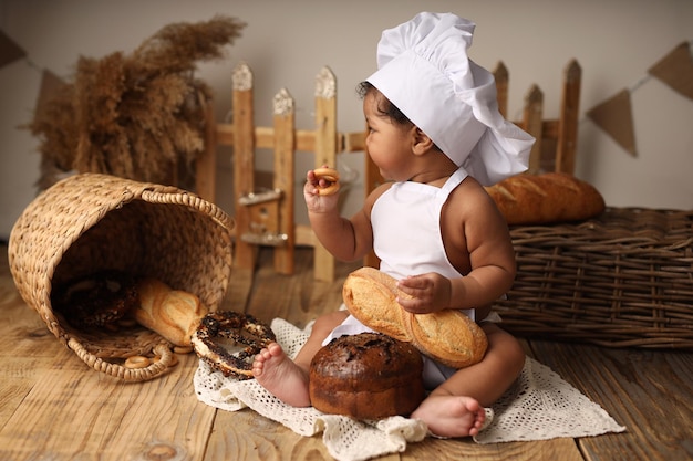 Uma criança fofa de pele escura com cabelo encaracolado e fantasiada de chef comendo um pãozinho e um pãozinho