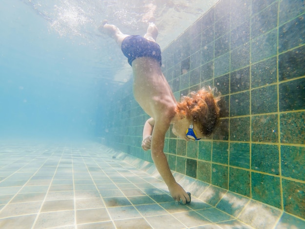 Uma criança menino está nadando debaixo d'água em uma piscina, sorrindo e prendendo a respiração, com óculos de natação