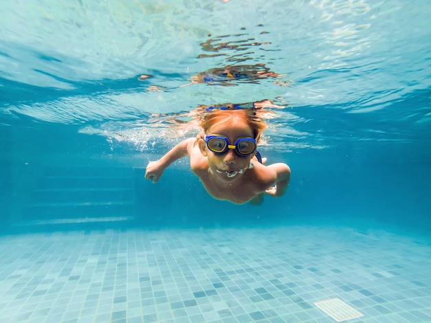 Uma criança menino está nadando debaixo d'água em uma piscina sorrindo e prendendo a respiração com óculos de natação