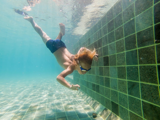Uma criança menino está nadando debaixo d'água em uma piscina, sorrindo e prendendo a respiração, com óculos de natação