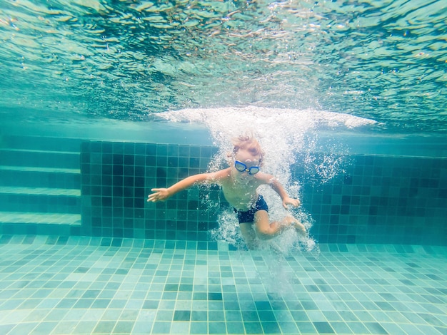 Uma criança menino está nadando debaixo d'água em uma piscina, sorrindo e prendendo a respiração, com óculos de natação