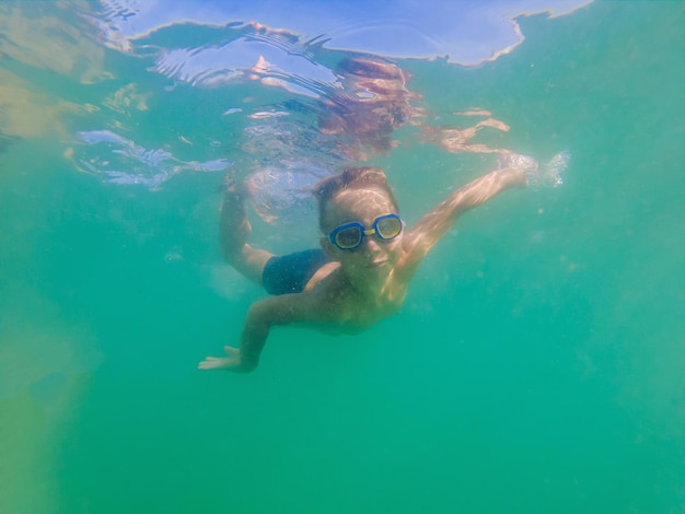 Uma criança menino está nadando debaixo d'água no mar, sorrindo e prendendo a respiração, com óculos de natação