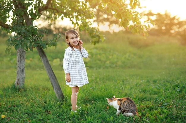 Uma criança no jardim está sorrindo docemente ao lado de um gato de rua ao pôr do sol Animais de estimação e seus donos