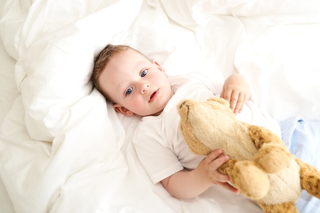 Uma criança pequena com olhos azuis segura um brinquedo deitado na cama. visão do topo.