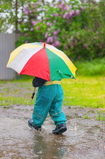 Uma criança pequena com um guarda-chuva colorido e brilhante caminha pelas poças na chuva