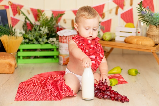 Uma criança pequena de um menino de seis meses está sentado no chão da cozinha com uma garrafa de leite e uvas