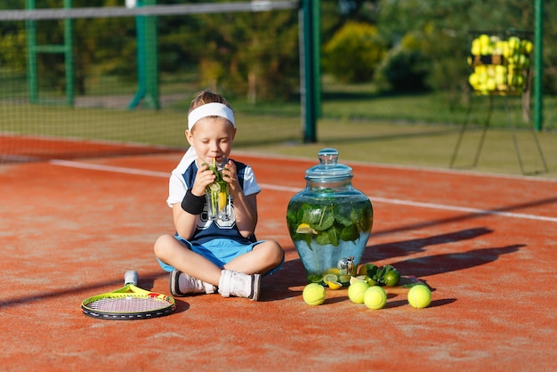 Uma criança pequena em um terno de tênis sentado em uma quadra de tênis no verão e bebendo limonada gelada Dispenser com uma bebida fresca e bolas de tênis no fundo